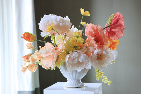 The image depicts a beautiful DIY faux floral centerpiece. The centerpiece consists of various faux flowers in vibrant peach and citrus hues, arranged in a decorative compote. Larger blooms including poppies and peonies serve as focal points.